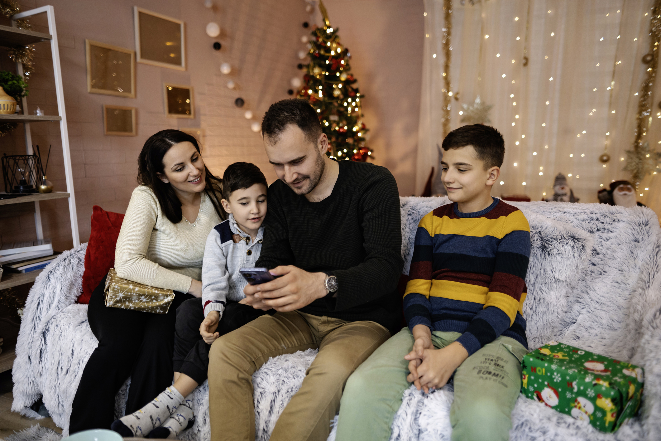 Изображение. Новогодний фон. Семья с детьми на диване смотрит на экран телефона, рассматривая цифровой подарок. Рядом лежат подарки традиционные в красивых упаковках