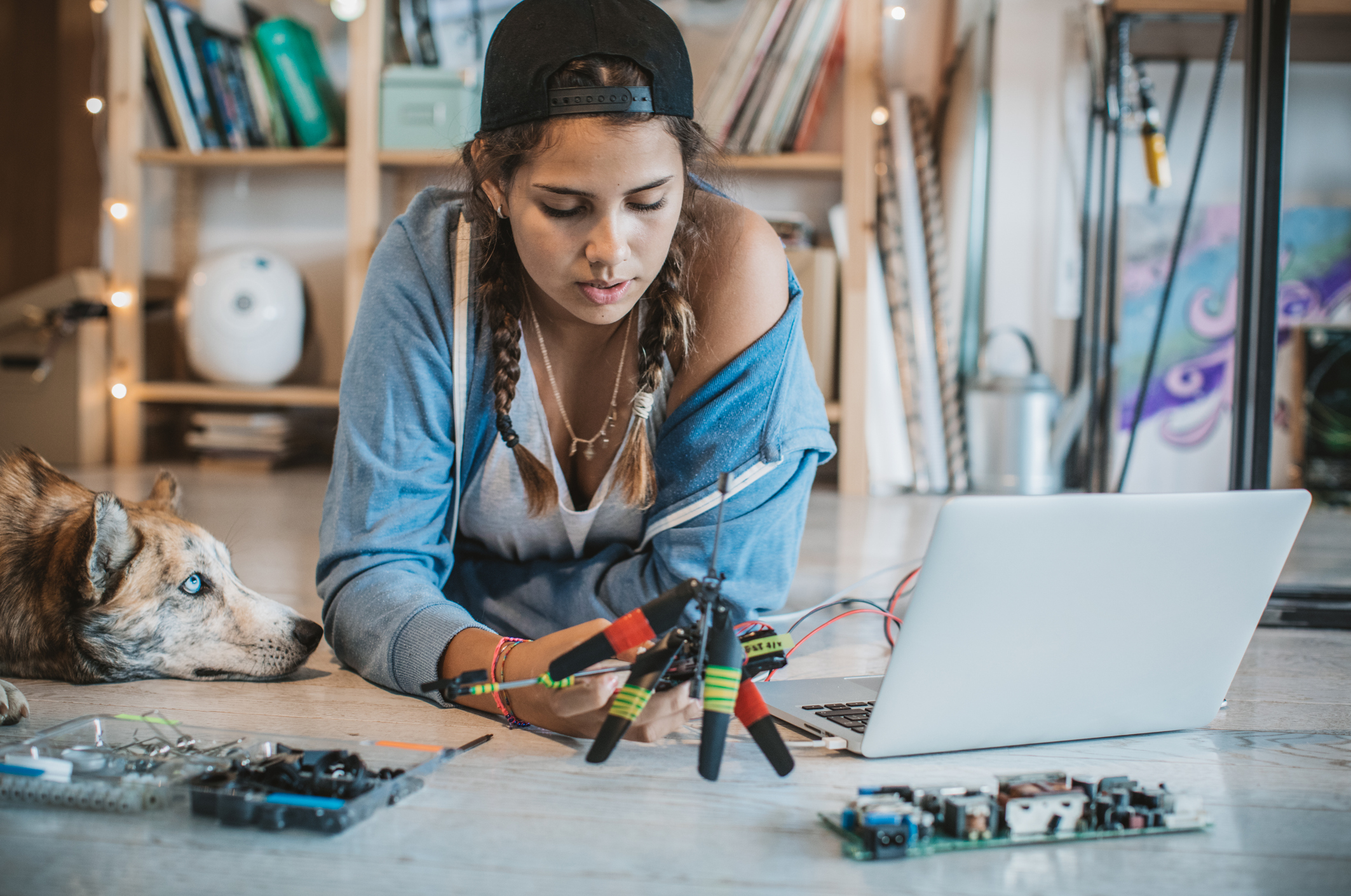Изображение. Девушка пытается собрать модель дрона за ноутбуком на полу, используя программу КОМПАС-3D Home для проектирования. Собака лежит рядом и смотрит на девушку